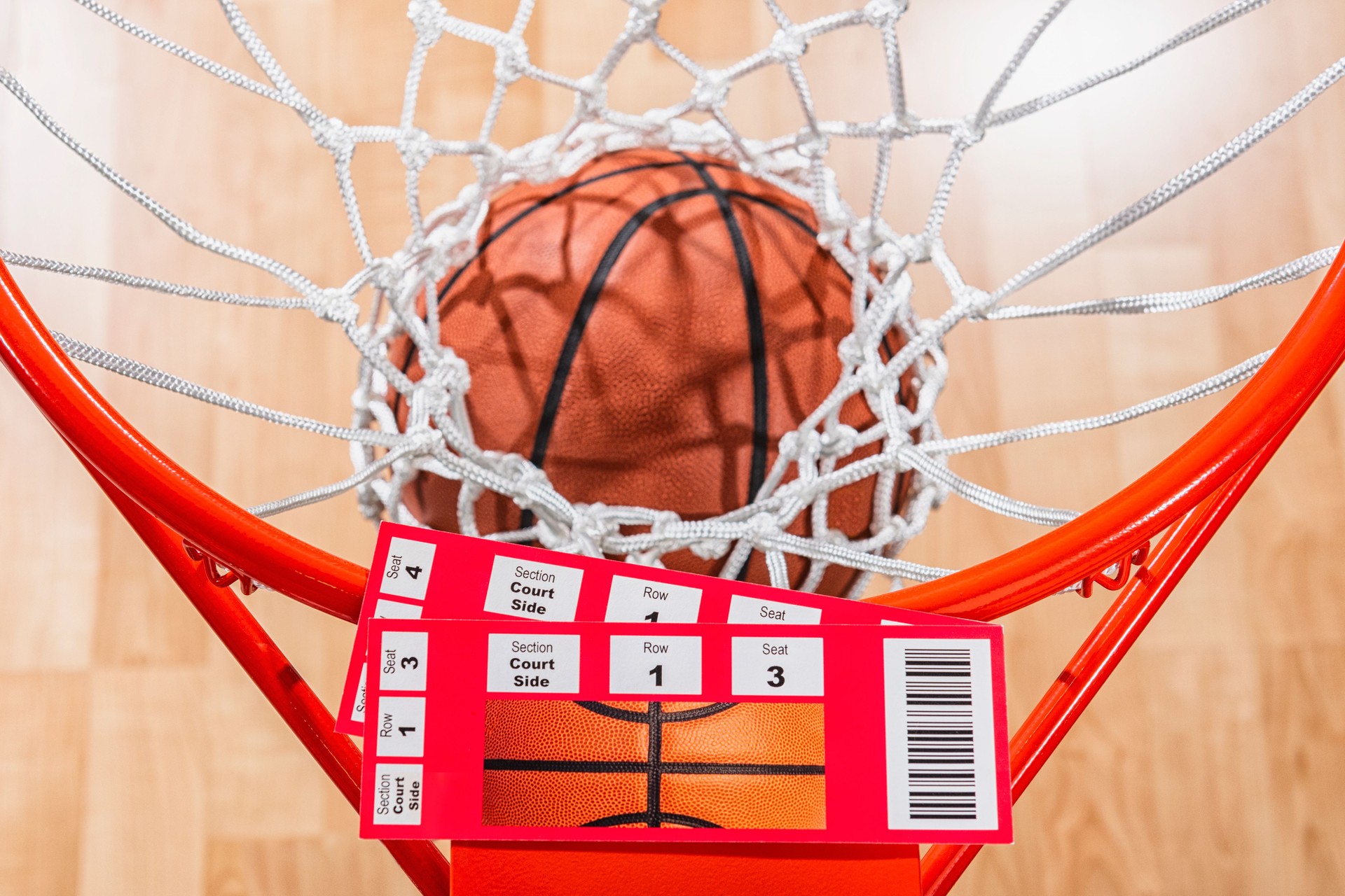 Looking down on a pair of basketball tickets as a ball goes through the net with arena wood floor in the background